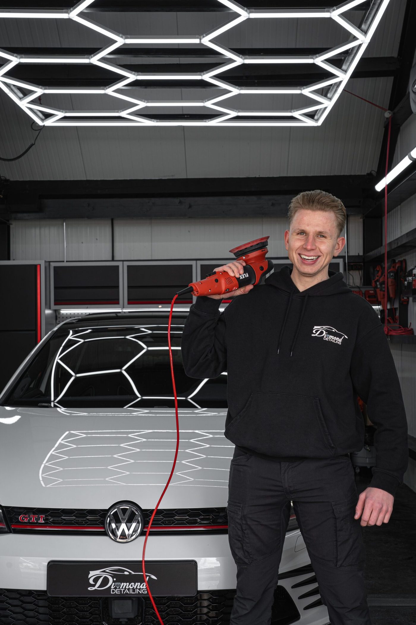 Danny Barten lacht naar de camera in zijn detailingstudio, polijstmachine op de schouder